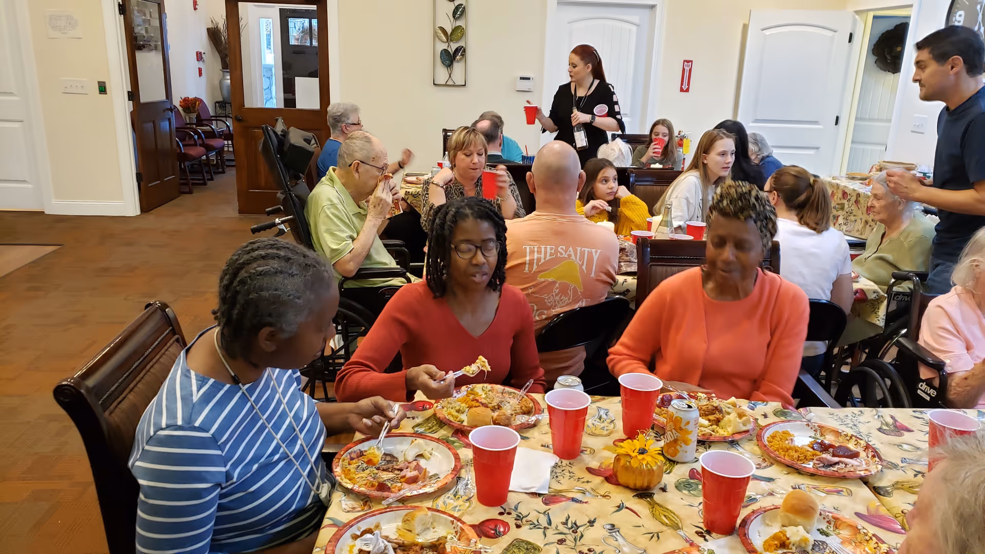 A group of elderly and middle-aged people sitting around tables in a dining area, eating food from plates and drinking from red cups. The room is bright with white walls and wooden doors, and there are decorations on the walls. Some people are engaged in conversation while others are eating.
