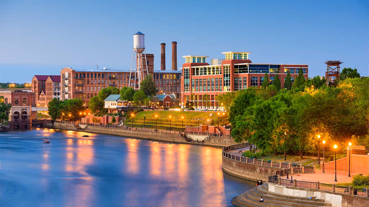 Dusk riverfront scene with brick buildings and a water tower, a lit promenade and trees reflecting on the water.