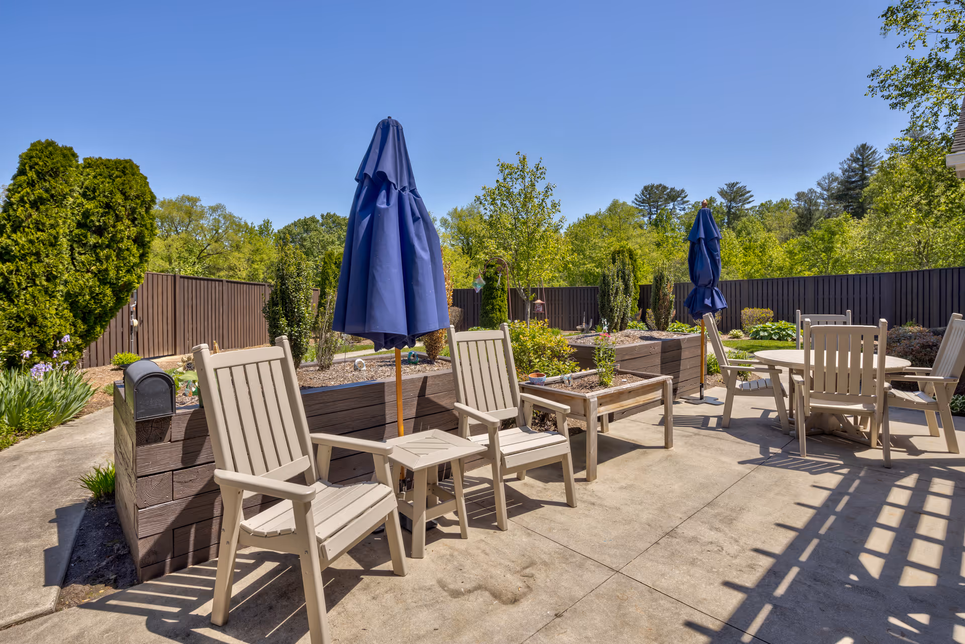 Outdoor patio area with beige chairs and tables, some with blue closed umbrellas, surrounded by raised garden beds and greenery under a clear blue sky.
