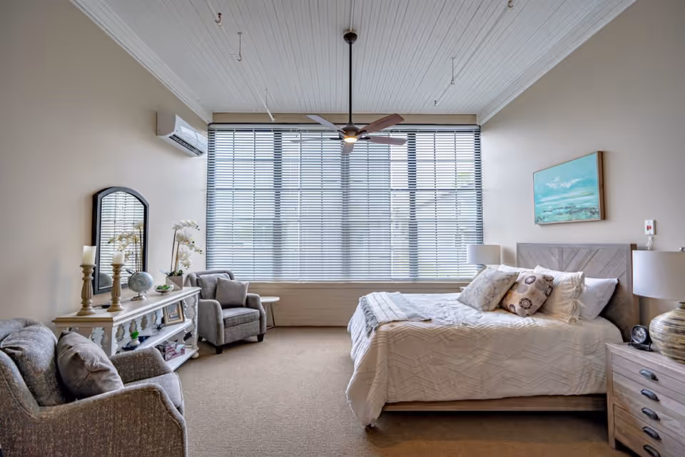 A bright and cozy bedroom with a large window covered by horizontal blinds. The room features a bed with white bedding and decorative pillows, two bedside tables with lamps, a comfortable armchair, a small sofa, and a white console table with candles, a globe, and a mirror. The walls are painted beige, and there is a ceiling fan with lights above the bed. A painting hangs on the wall above the bed.