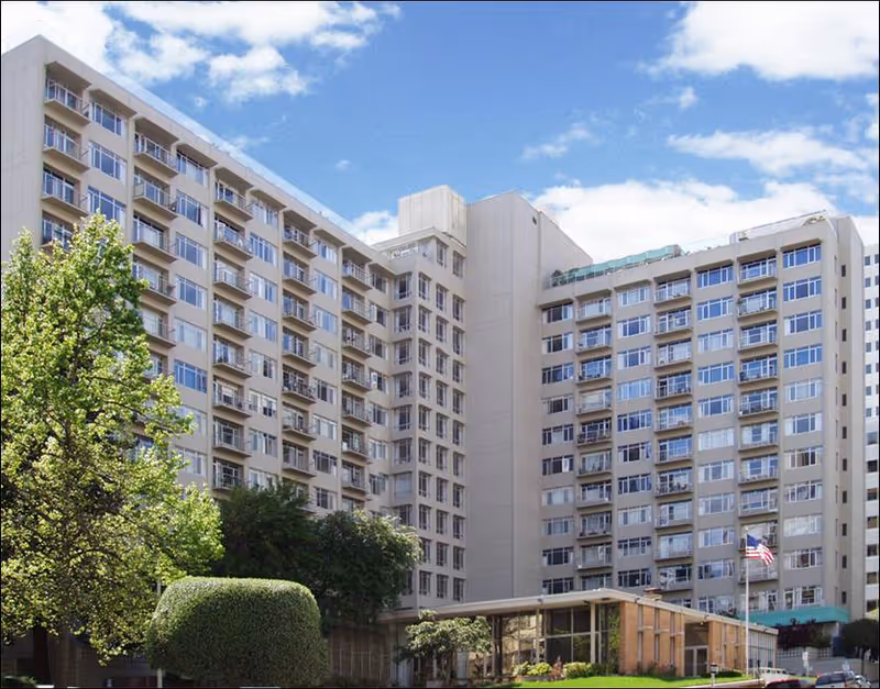 Exterior view of a multi-story senior living facility building with many windows and balconies, surrounded by trees and greenery under a partly cloudy sky.