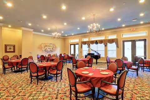 Spacious dining room with round tables draped in red tablecloths, upholstered chairs, chandeliers, and a grand piano by large windows.
