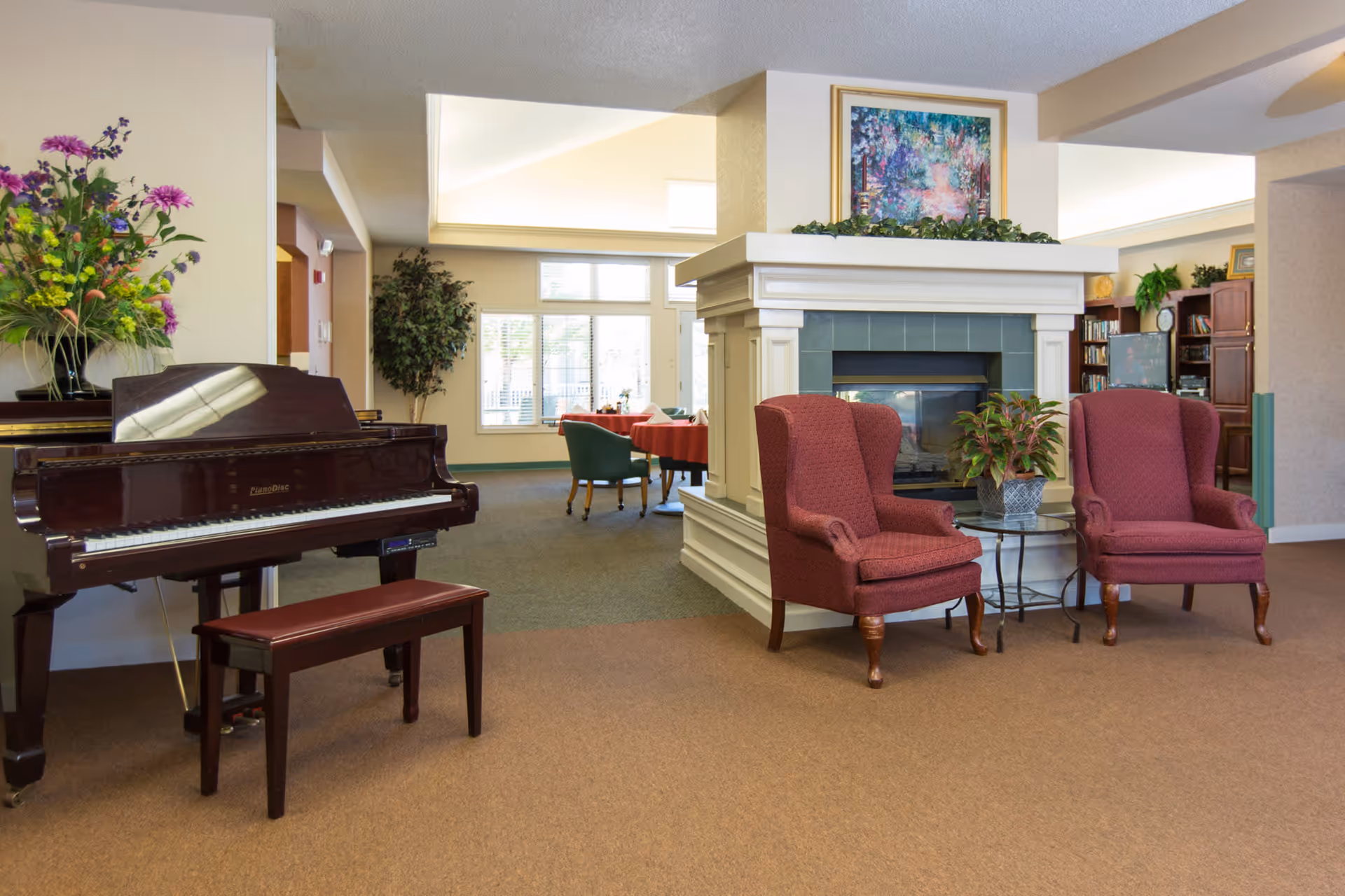 A cozy senior living facility common area with a dark wood piano and matching bench on the left, a large floral arrangement on top of the piano, two red upholstered armchairs with wooden legs positioned around a small glass-top table with a potted plant, and a white fireplace with green tiles and a colorful framed painting above it. In the background, there is a dining area with tables covered in red tablecloths and green chairs, large windows letting in natural light, and bookshelves with a TV.