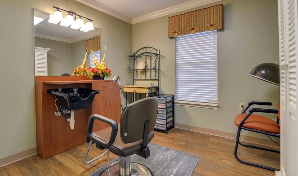 Interior view of a senior living facility hair salon area featuring a black salon chair in front of a wooden counter with a black wash basin. There is a large mirror above the counter with a light fixture, a floral arrangement on the counter, a window with blinds and a plaid valance, a metal shelving unit with a small desk, and two orange chairs with black armrests along the wall.