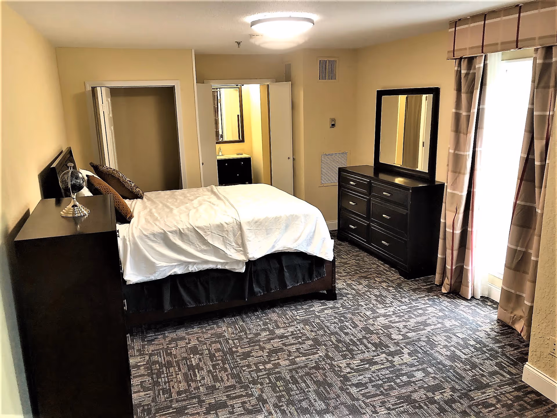 A senior living community bedroom with a bed covered in white bedding and brown pillows, a dark wood dresser with a mirror, a matching nightstand with a decorative globe, patterned carpet flooring, beige walls, and a window with beige and brown plaid curtains letting in natural light.