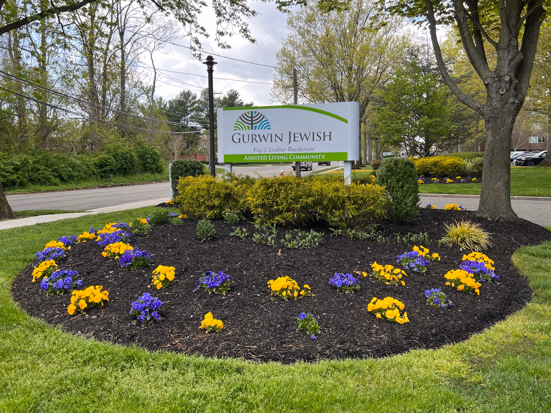 Entrance sign reading 'Gurwin Jewish Fay J. Lindner Residences' surrounded by a landscaped bed of yellow and purple flowers and trees.