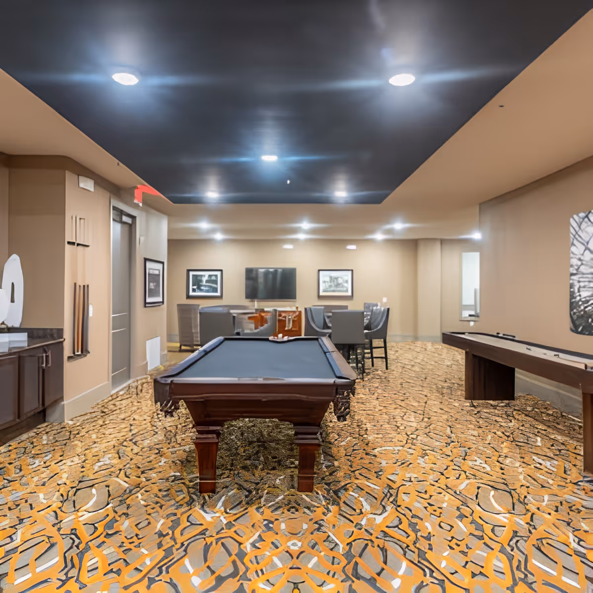 A recreational room featuring a pool table in the center, a shuffleboard table to the right, and a seating area with chairs and a TV in the background. The room has a patterned carpet, beige walls, and a dark ceiling with recessed lighting.