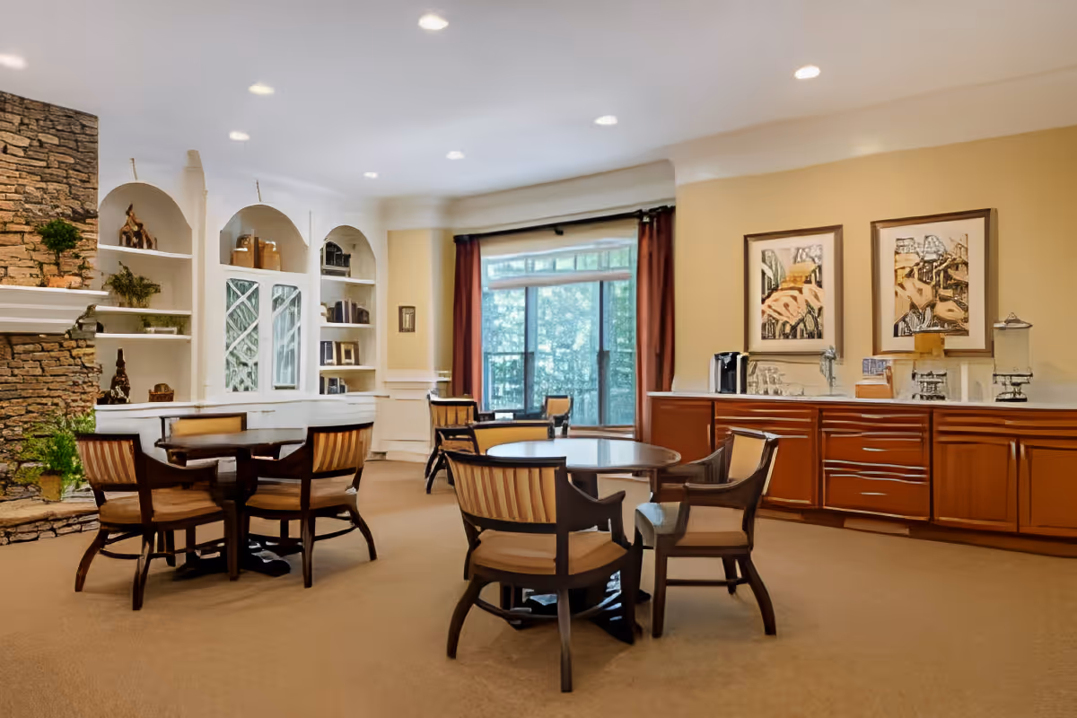 A cozy senior living common area with round wooden tables and cushioned chairs arranged on a carpeted floor. The room features a stone fireplace on the left, built-in white shelves with decorative items, large windows with red curtains letting in natural light, and a wooden cabinet with a coffee station and framed artwork above it on the right wall.