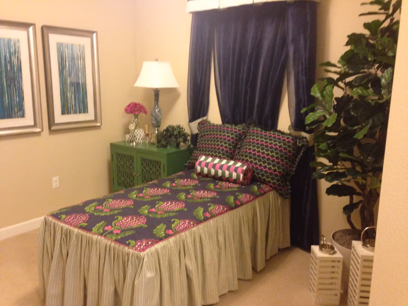 A small bedroom with a single bed covered in a floral and patterned bedspread with matching pillows. There is a green nightstand with a lamp, flowers, and decorative items next to the bed. Two framed abstract artworks hang on the wall, and a large potted plant is placed in the corner near the window with dark curtains.
