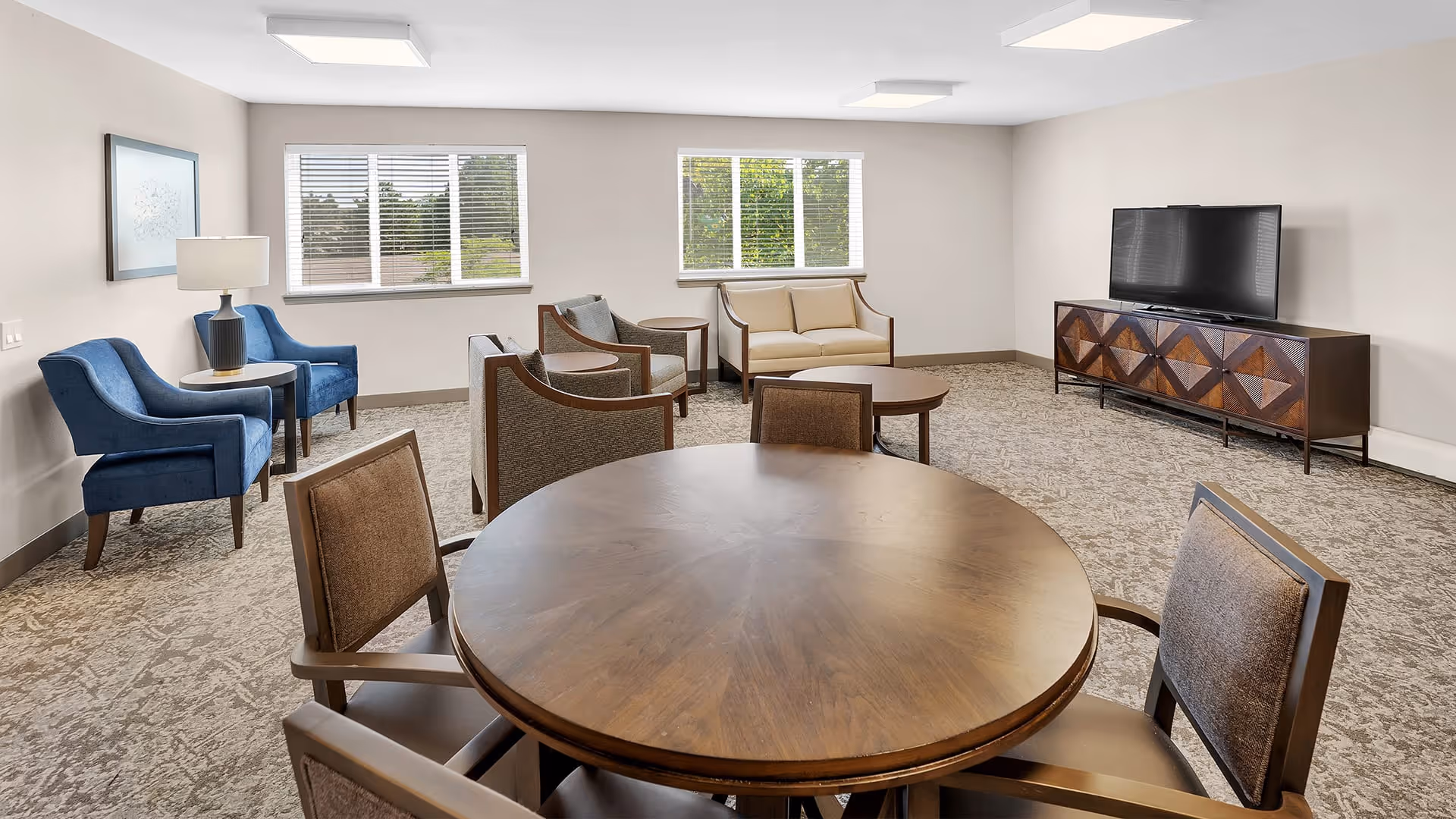 A spacious living room with a round wooden table and four chairs in the foreground. The room features two blue armchairs with a small round table and lamp between them on the left side, a beige loveseat and two brown armchairs arranged around a round coffee table near the windows. A large flat-screen TV sits on a wooden media console against the right wall. The room has carpeted flooring, white walls, and two windows with blinds letting in natural light.