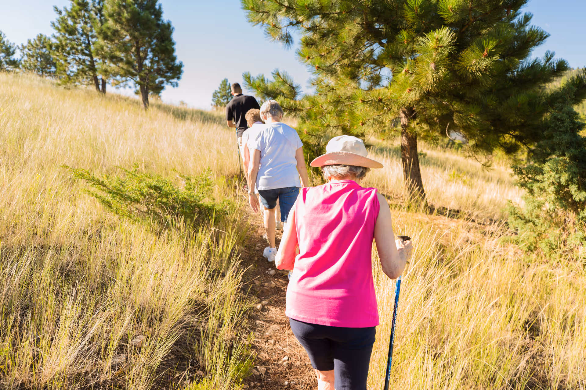 A group of four people walking up a narrow dirt path through a grassy area with trees on a sunny day. The person closest to the camera is an older woman wearing a pink sleeveless top and a wide-brimmed hat, using a walking stick.