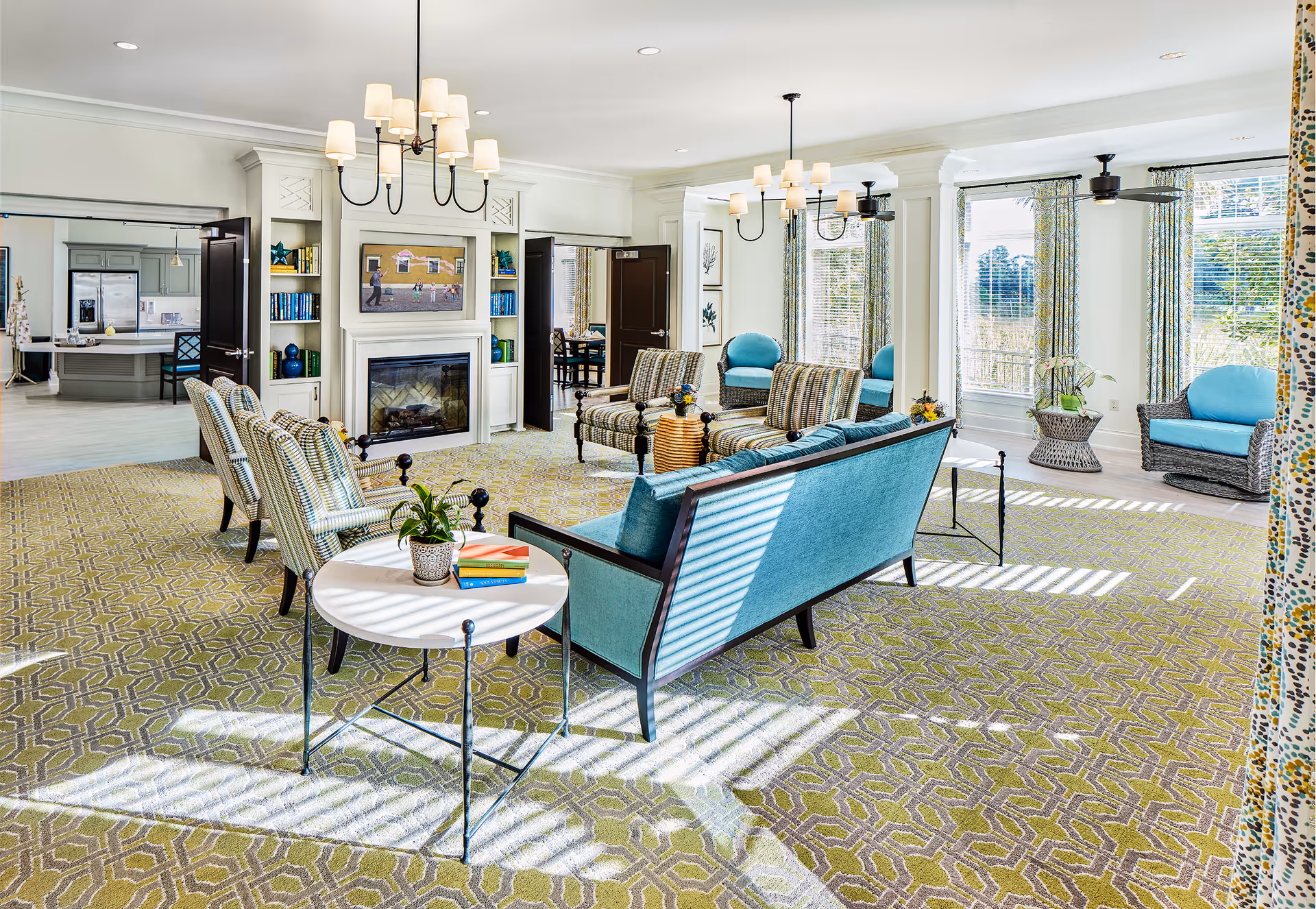 A bright and spacious living room area with patterned carpet, multiple seating options including a teal sofa, striped armchairs, and wicker chairs with teal cushions. The room features large windows with patterned curtains allowing natural light to stream in, a fireplace with built-in shelves on either side, and two chandeliers hanging from the ceiling. In the background, a kitchen area is visible through open double doors.