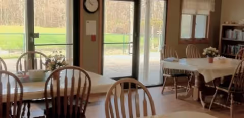 Sunlit dining room with round tables and wooden chairs set with tablecloths and small flower centerpieces, looking out through glass doors to a grassy outdoor area.