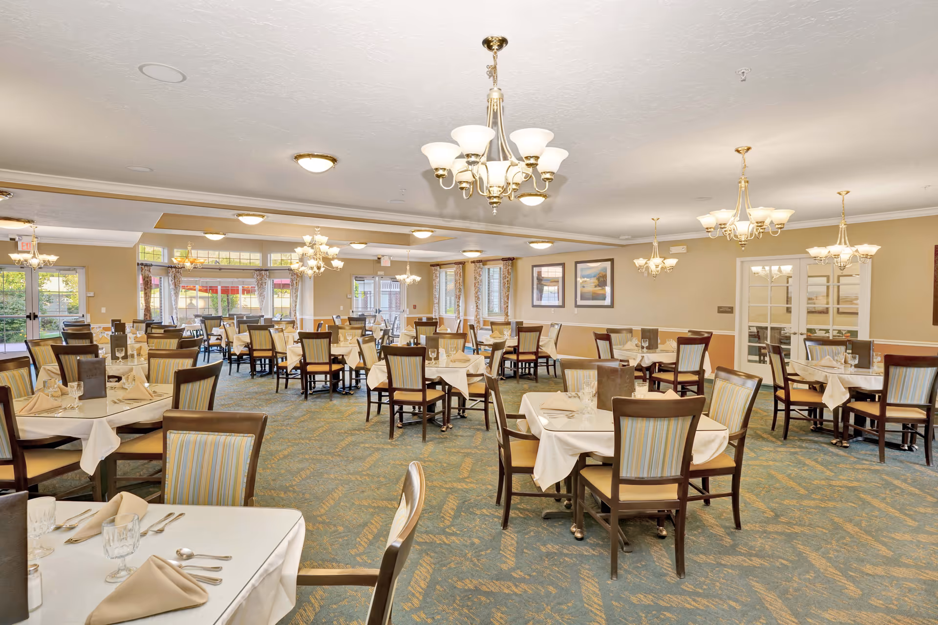 A spacious dining room with multiple tables covered in white tablecloths, each set with beige napkins, glassware, and silverware. The room features patterned carpet, several chandeliers hanging from the ceiling, large windows with curtains allowing natural light, and framed artwork on the walls.