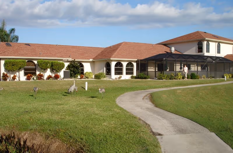 A one-story building with a red roof and screened porch sits behind a curved concrete walkway across a grassy lawn where several large birds graze.