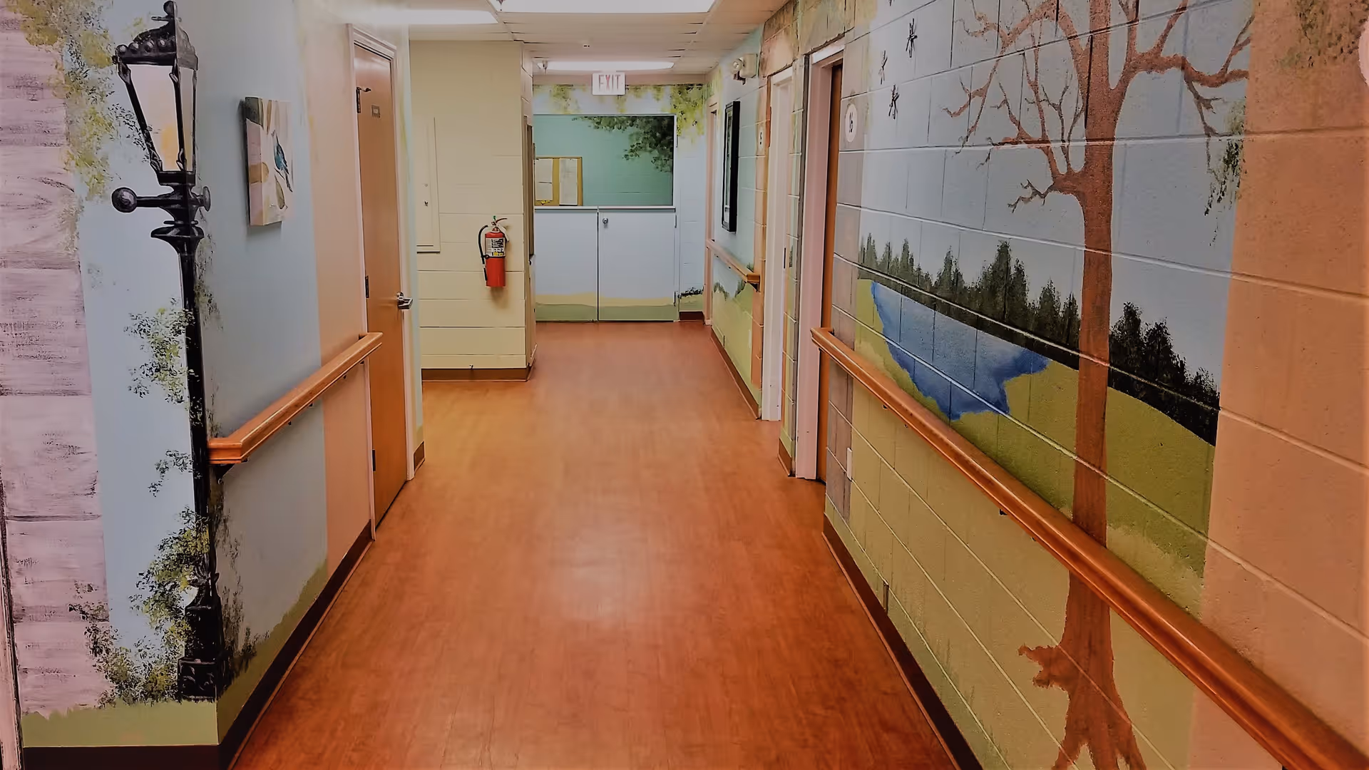 A hallway in a senior living facility with wooden flooring and handrails on both sides. The walls are decorated with murals depicting outdoor scenes including trees, a pond, and a lamppost. There are several doors along the hallway and an exit sign at the far end.