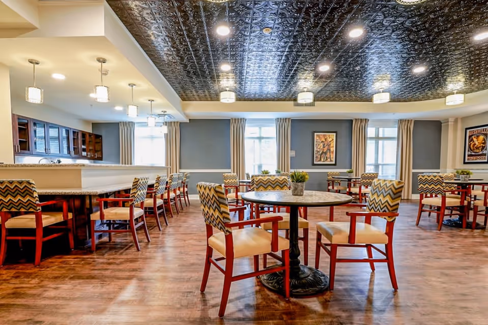 A spacious dining room with multiple round and rectangular tables surrounded by chairs with patterned backs and cushioned seats. The room features a decorative dark ceiling with recessed lighting and pendant lights over a counter area. Large windows with curtains allow natural light to fill the space, and framed artwork is displayed on the walls.