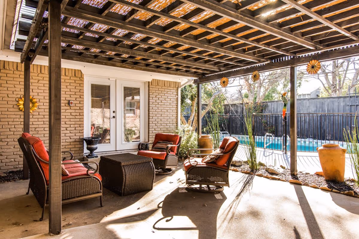 Outdoor covered patio area with wicker furniture including a loveseat and two chairs with red cushions, a wicker coffee table, and decorative sunflowers hanging from the wooden pergola. Beyond the patio is a fenced swimming pool surrounded by trees and plants.
