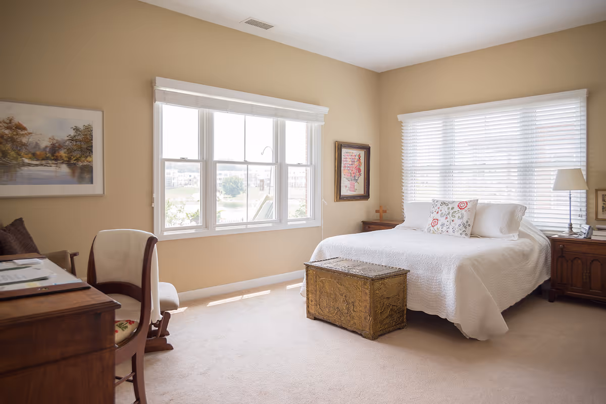Sunny furnished bedroom with a white bed, wooden nightstands, a decorative trunk at the foot and large windows letting in natural light.