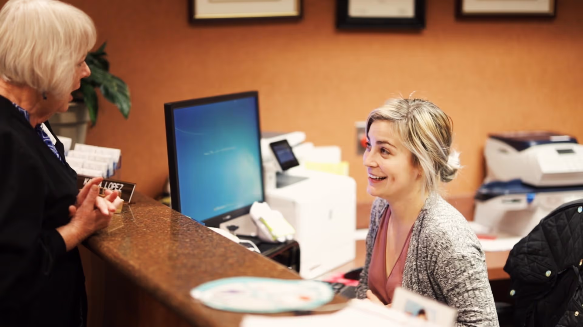 A receptionist with blonde hair tied back and wearing a gray cardigan smiles and talks to an elderly woman with short white hair standing at the reception desk. The desk has a computer monitor, office equipment, and a nameplate that reads 'LOPEZ'. The background shows a warm-colored wall with framed certificates or documents.