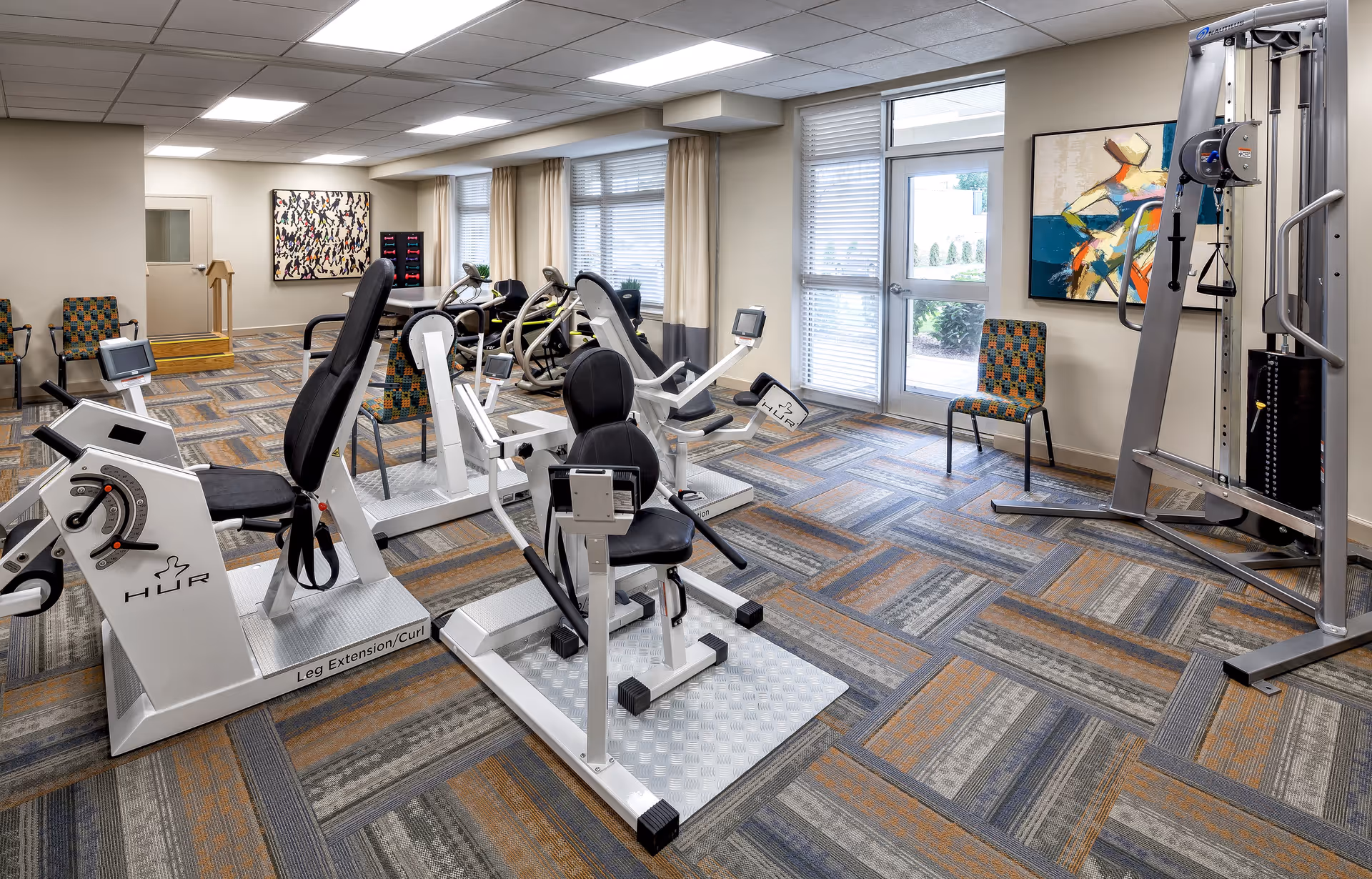 A well-lit fitness room with various exercise machines including leg extension/curl equipment, a multi-functional weight machine, and several chairs along the walls. The room has large windows with beige curtains, patterned carpet flooring, and colorful abstract paintings on the walls.