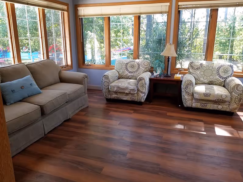 A bright living room with large windows showing outdoor greenery. The room has a beige sofa with a blue pillow on the left and two patterned armchairs on the right, separated by a wooden side table with a lamp and some small items. The floor is wooden with a warm tone.