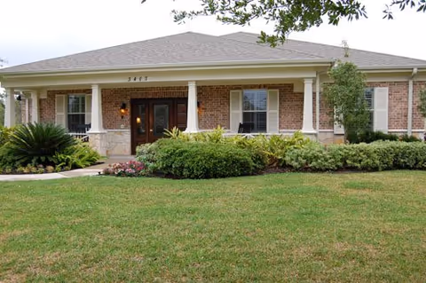 Single-story brick building with a covered front porch, white columns, and a manicured lawn and shrubs.