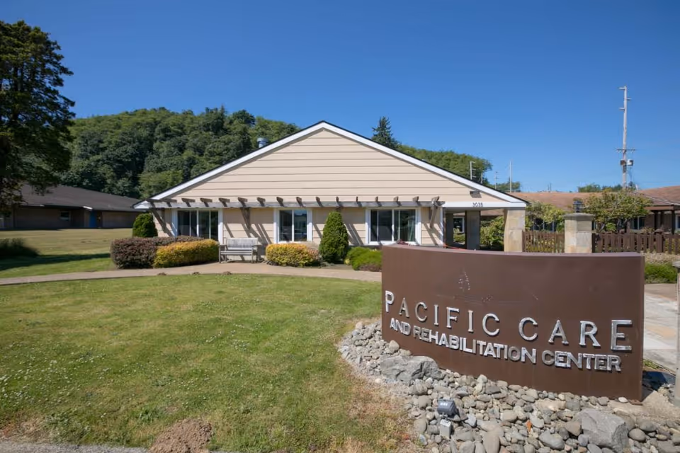 Exterior view of Pacific Care and Rehabilitation Center building with a large brown sign in front on a grassy lawn, surrounded by bushes and trees under a clear blue sky.