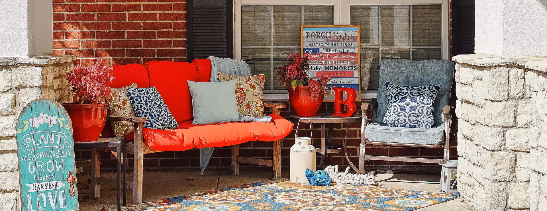A cozy porch seating area with a bright red cushioned bench and a blue cushioned chair, both adorned with decorative pillows. There are potted plants, a colorful patterned rug, and various decorative signs including one that says 'Welcome' and another with porch rules. The porch is framed by stone pillars and a brick wall with a window in the background.