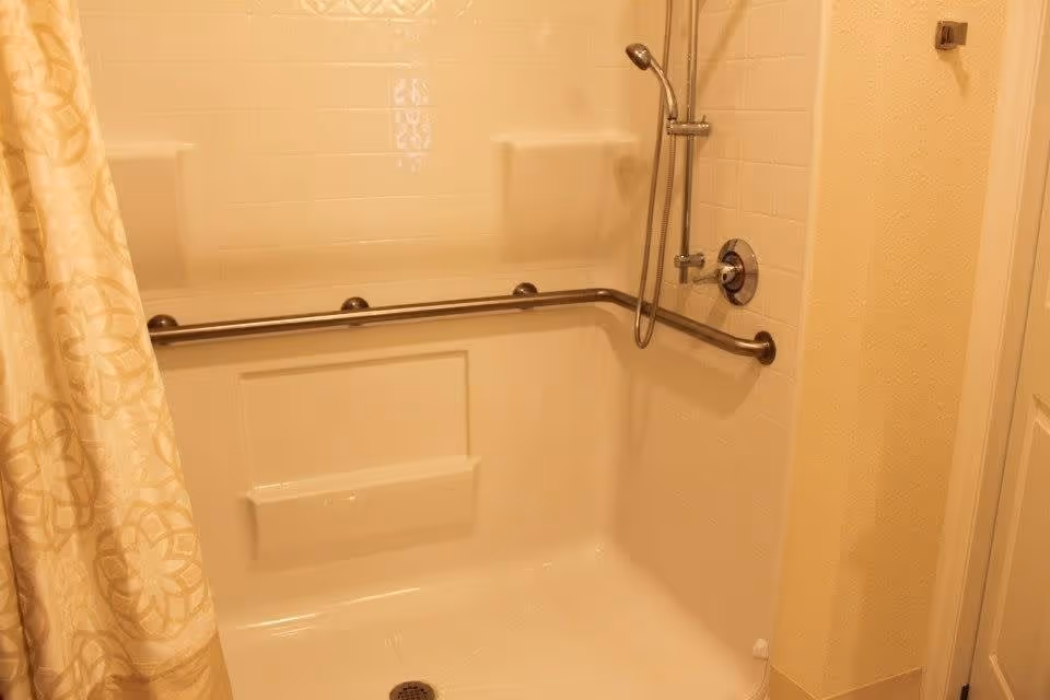 A beige tiled shower area with a built-in seat and a horizontal grab bar along the back and side walls. There is a handheld showerhead attached to a vertical bar on the right wall. A patterned beige and white shower curtain is partially visible on the left side.