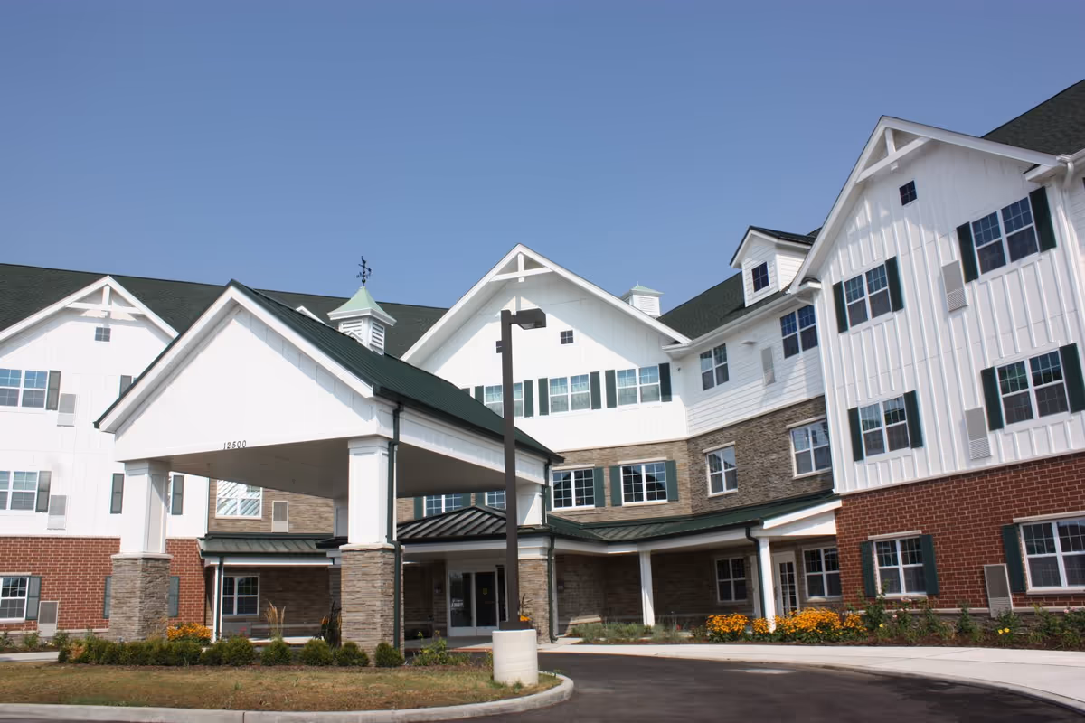 Exterior view of a multi-story senior living facility building with white siding, brick and stone accents, green shutters, and a covered entrance with pillars. The sky is clear and blue, and there are landscaped flower beds near the entrance.