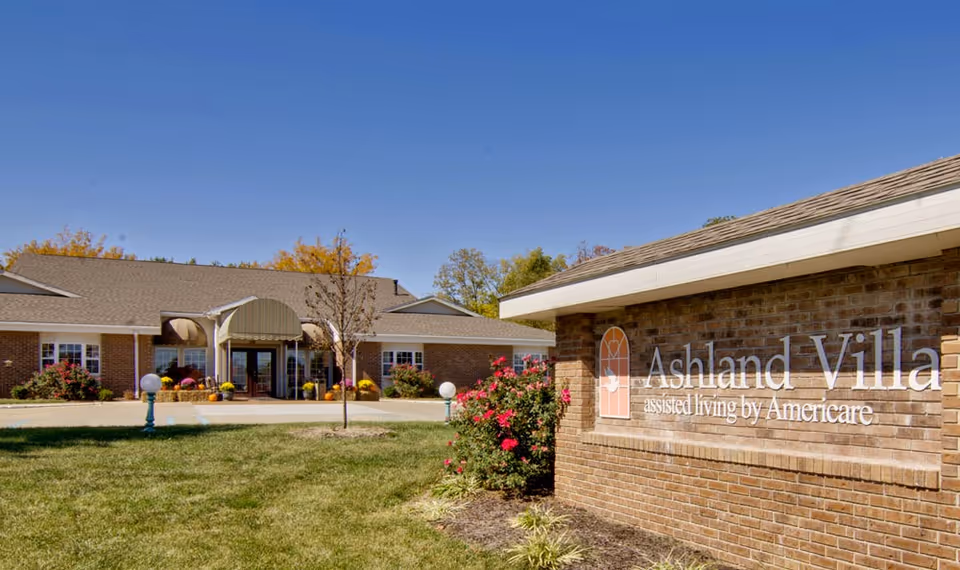 Exterior view of Ashland Villa assisted living facility on a clear sunny day, showing a brick building with a covered entrance, landscaping with bushes and flowers, and a sign with the facility name on a brick wall in the foreground.