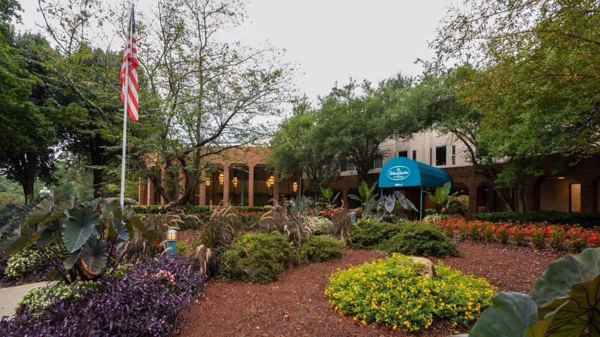 Exterior view of Delmar Gardens of Smyrna senior living facility showing a landscaped garden with various plants and flowers, an American flag on a flagpole, and a building entrance with a blue awning displaying the facility's name.