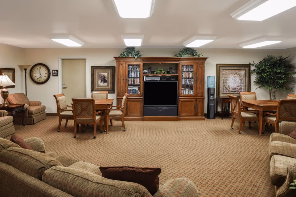 Furnished common room with sofas, tables, chairs, an entertainment center and TV in a senior living facility.