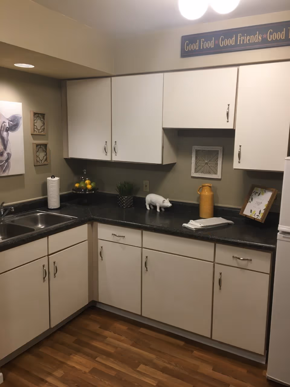 A small kitchen area with white cabinets, a black countertop, and a double sink. On the countertop are a paper towel roll, a tiered fruit stand with lemons, a small potted plant, a white pig figurine, a yellow pitcher, folded white towels, and a framed sign. The walls are beige with a cow painting and decorative wall hangings. Above the cabinets is a sign that reads 'Good Food • Good Friends • Good Times'. The floor is wooden.