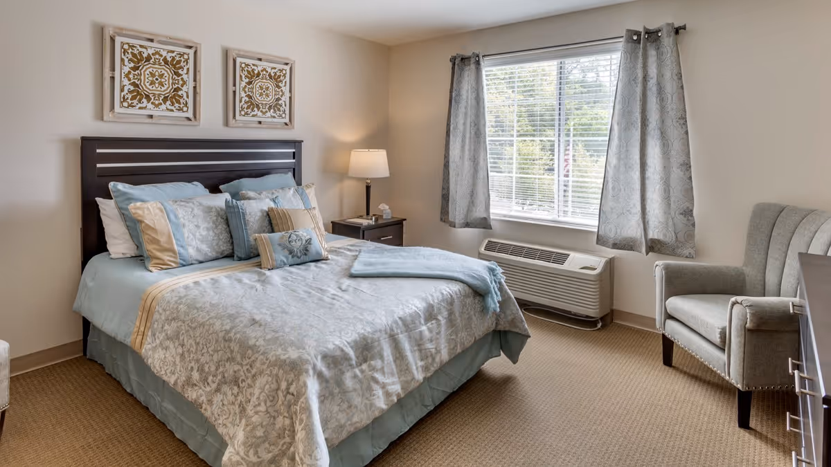 A neatly made bed with multiple pillows and a light blue and beige patterned comforter in a senior living facility bedroom. The room has beige walls, a window with gray patterned curtains, a nightstand with a lamp and tissue box, a gray upholstered armchair, and a heating/cooling unit beneath the window.