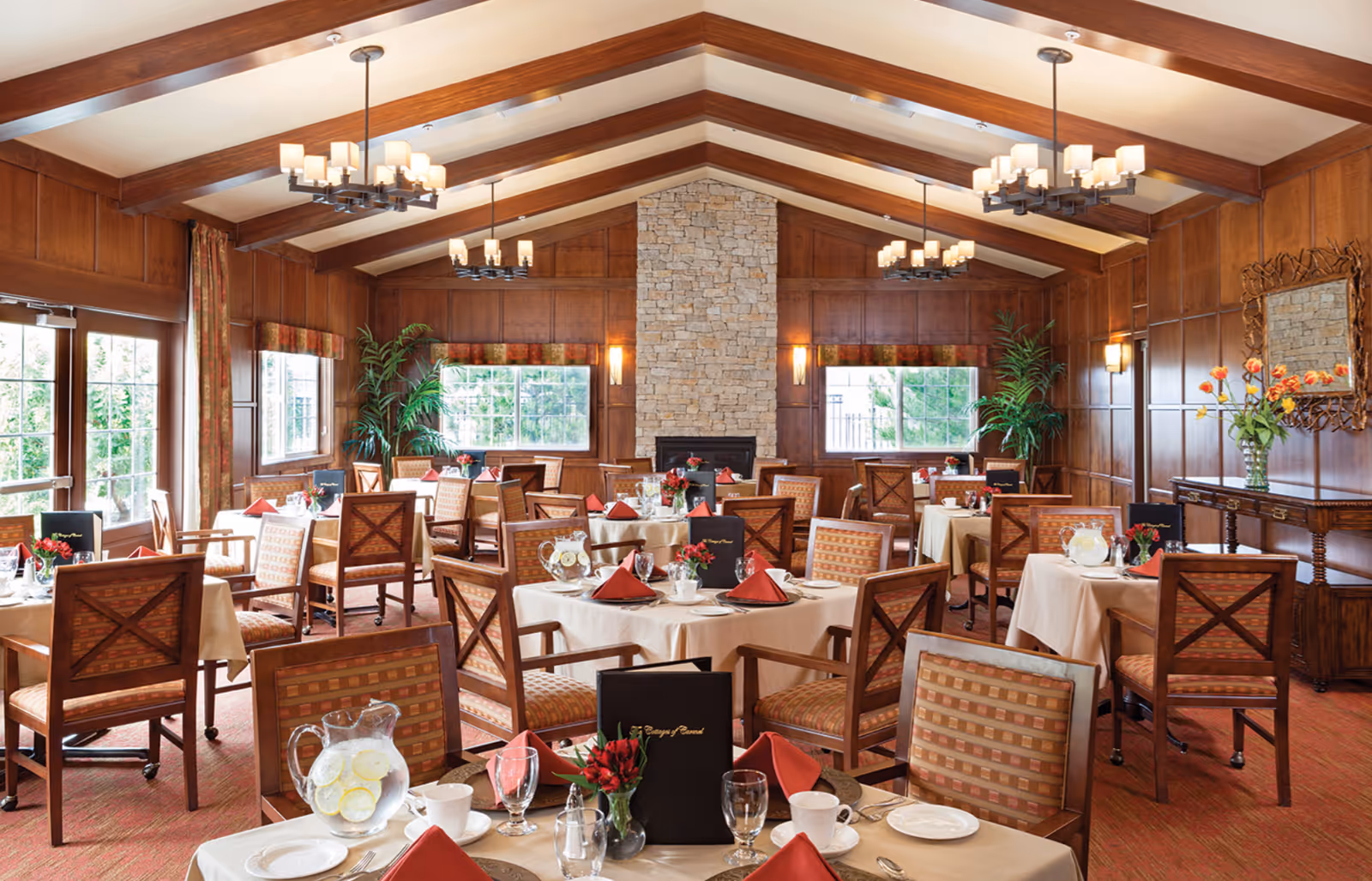 A warmly lit dining room with wooden panel walls and exposed wooden beams on the ceiling. Multiple round tables are set with white tablecloths, red napkins, glassware, and small flower arrangements. There is a stone fireplace centered on the far wall, flanked by windows with views of greenery outside. The room features elegant chandeliers, potted plants, and a decorative mirror above a wooden sideboard with a vase of flowers.