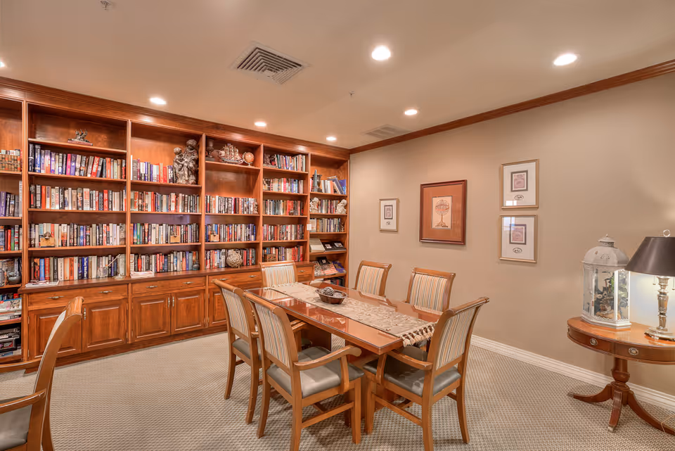 A cozy interior room featuring a wooden table with six cushioned chairs arranged around it. Behind the table is a large built-in wooden bookshelf filled with books and decorative items. The walls are beige with framed artwork, and there is a small round side table with a lamp and decorative lantern on it. The room is well-lit with recessed ceiling lights.