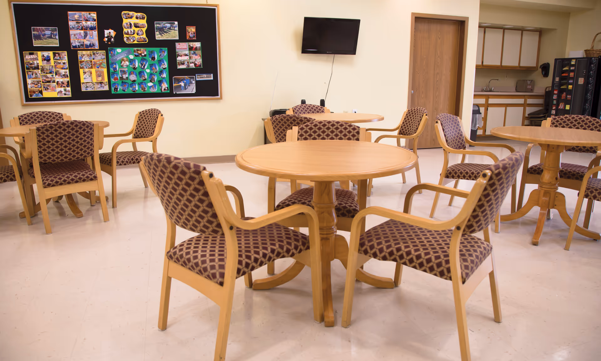 Empty senior living common room with round wooden tables and patterned chairs, a bulletin board and wall-mounted TV.