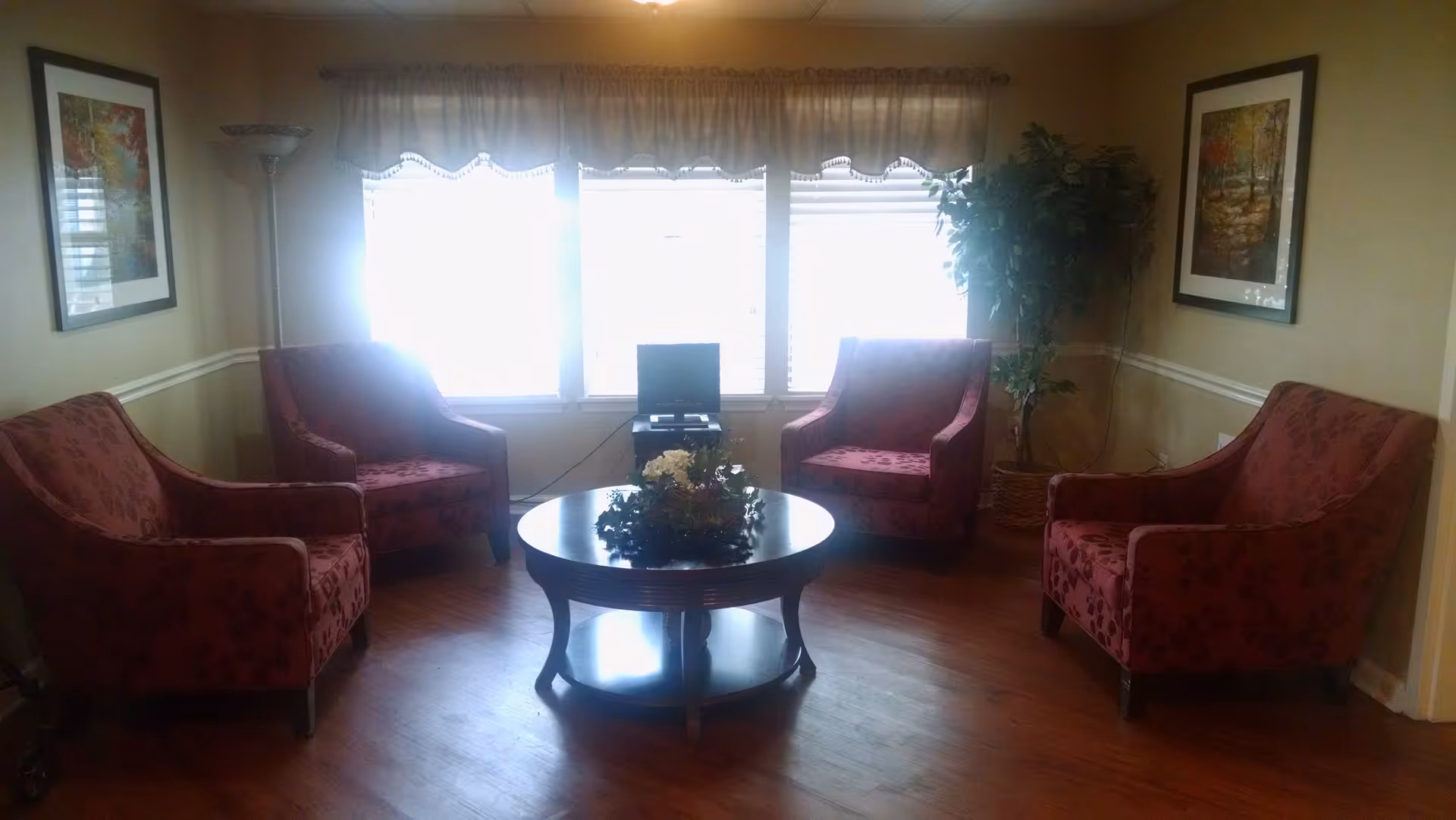 A cozy living room area with four red patterned armchairs arranged around a round wooden coffee table with a floral centerpiece. Behind the chairs are three large windows with sheer curtains, a small TV on a stand, a tall floor lamp, two framed landscape paintings on the walls, and a potted plant in the corner.