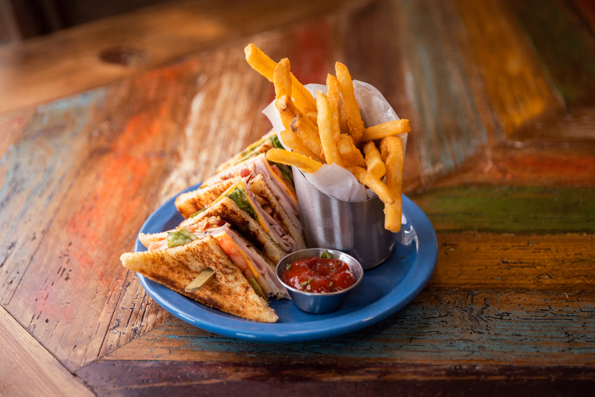 A blue plate with a club sandwich cut into four triangular pieces, a metal cup filled with French fries, and a small metal container of ketchup on a rustic wooden table.