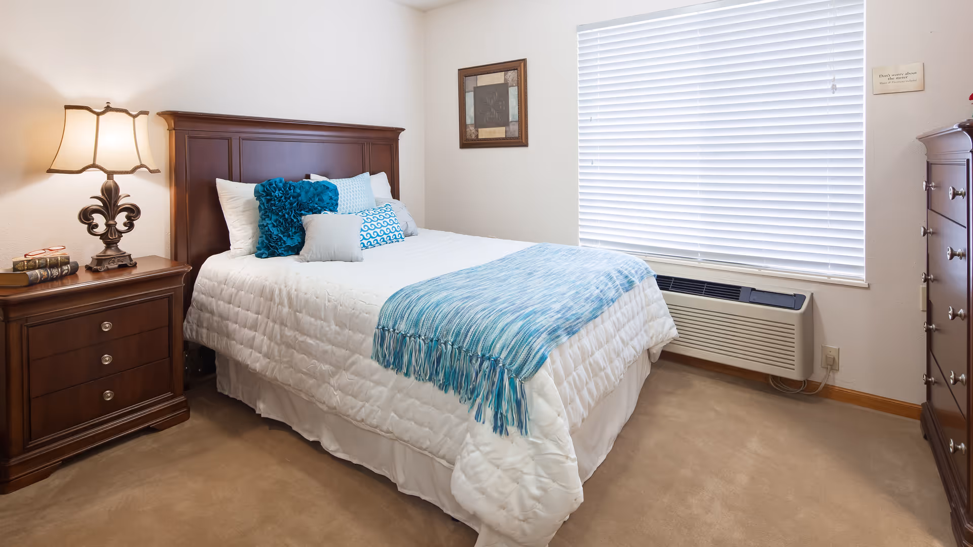 A neatly made bed with white bedding and blue accent pillows and throw blanket in a bedroom. The room has beige carpet, a wooden nightstand with a lamp and books, a wooden dresser, a window with closed blinds, and a framed picture on the wall.