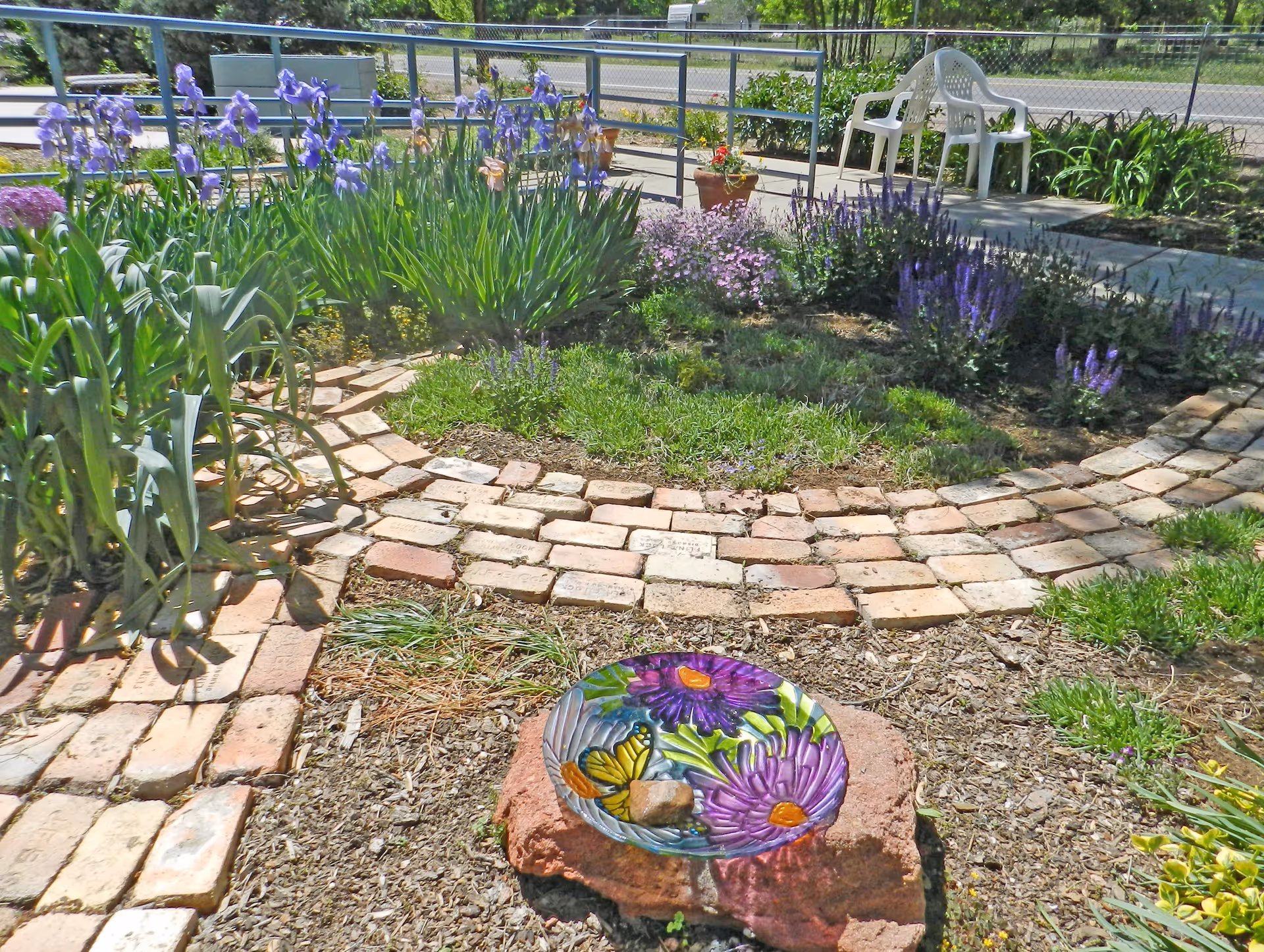 A garden area with a circular brick pathway surrounding a patch of grass and various flowering plants including purple irises and other purple flowers. In the foreground, there is a decorative glass bowl with painted flowers and a butterfly, placed on a rock. A white plastic chair is visible near a metal railing in the background.