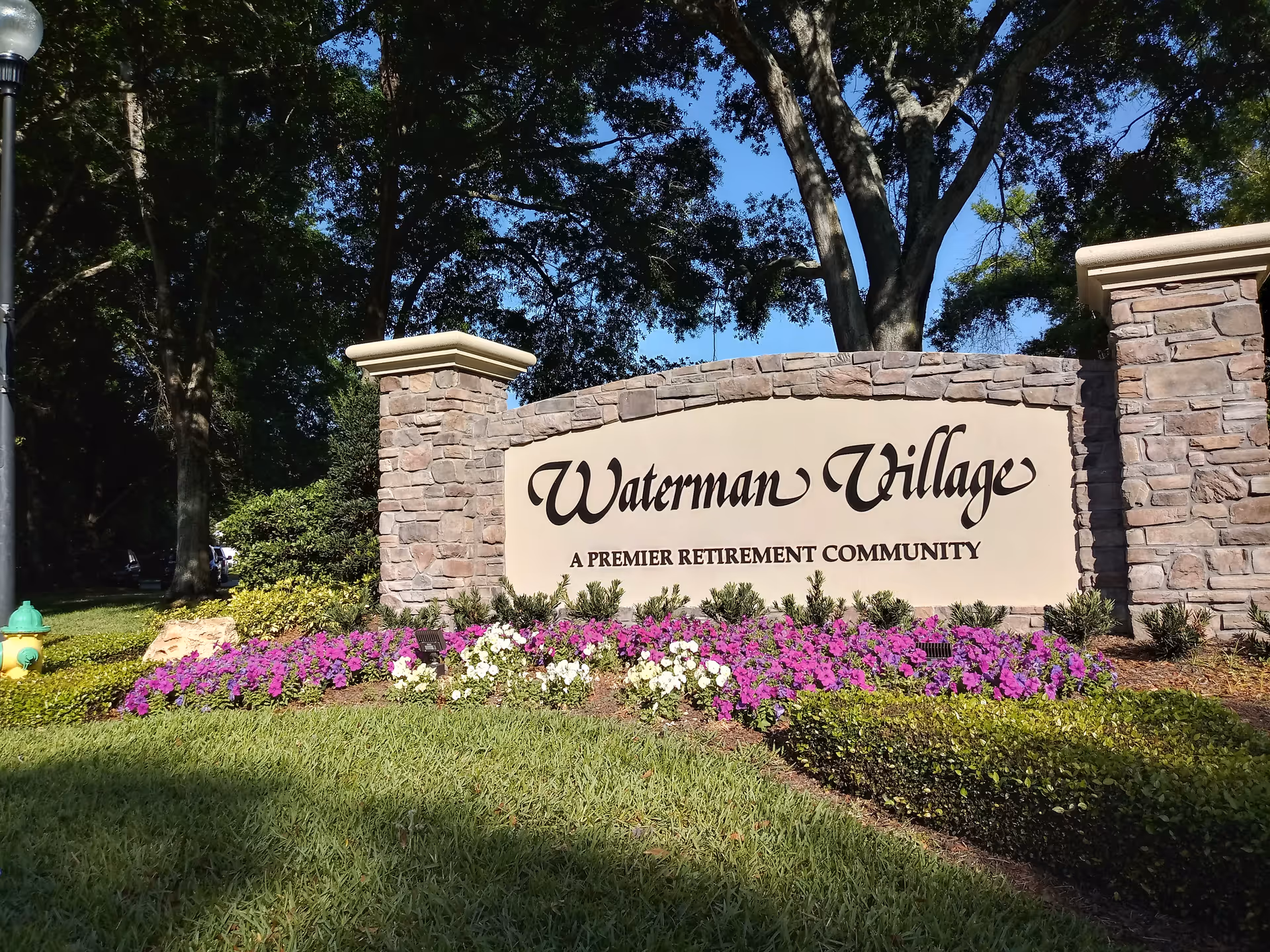 Stone entrance sign for Waterman Village, a premier retirement community, surrounded by green grass, colorful flowers, and trees in the background under a clear blue sky.