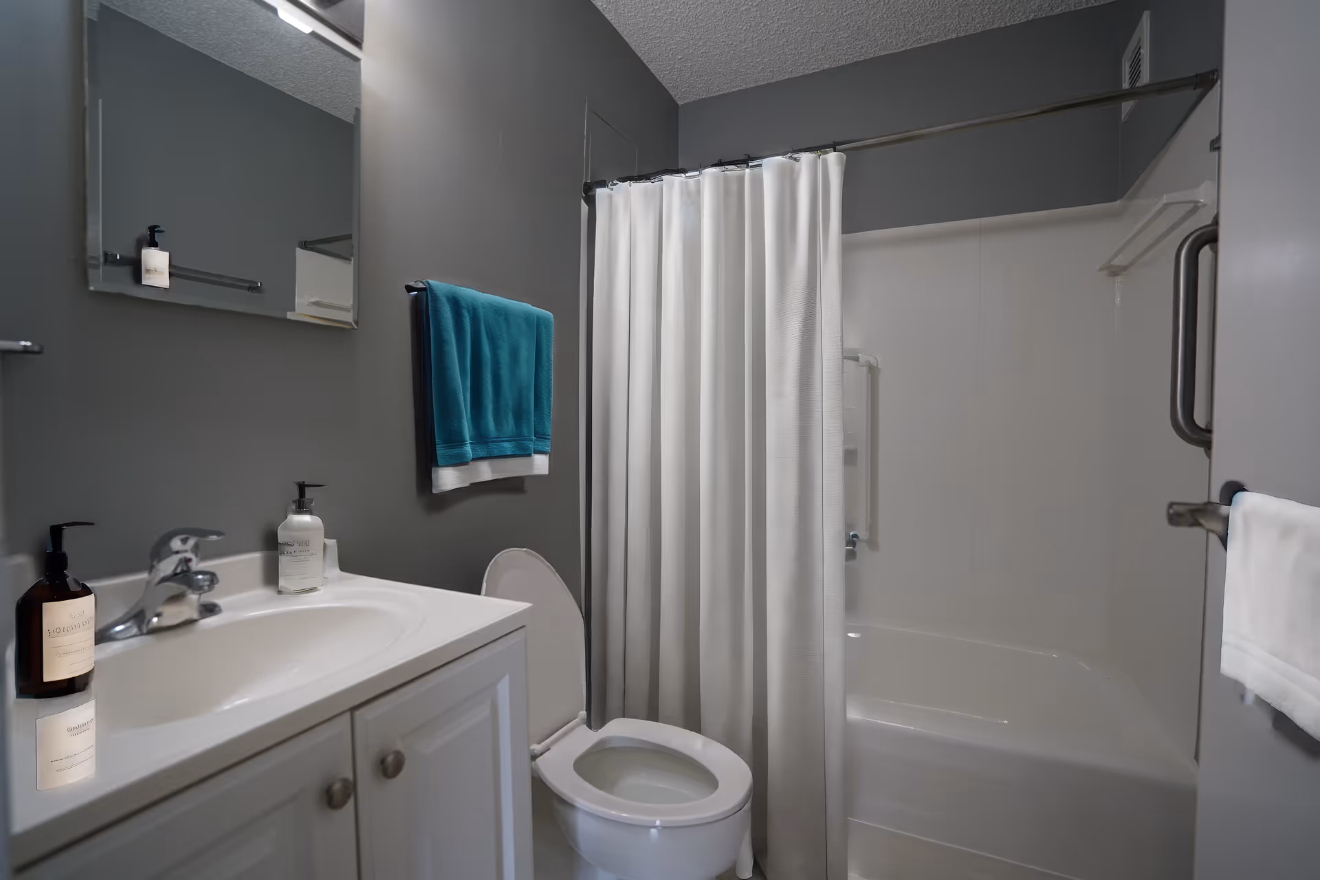 A clean bathroom with gray walls featuring a white sink cabinet with a faucet and soap dispensers, a toilet with the lid open, a white bathtub with a white shower curtain, and a teal towel hanging on the wall.