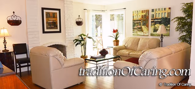 A cozy living room with beige leather sofas arranged around a glass coffee table. The room has wooden flooring, a white brick fireplace, framed artwork on the walls, potted plants, and large windows letting in natural light.