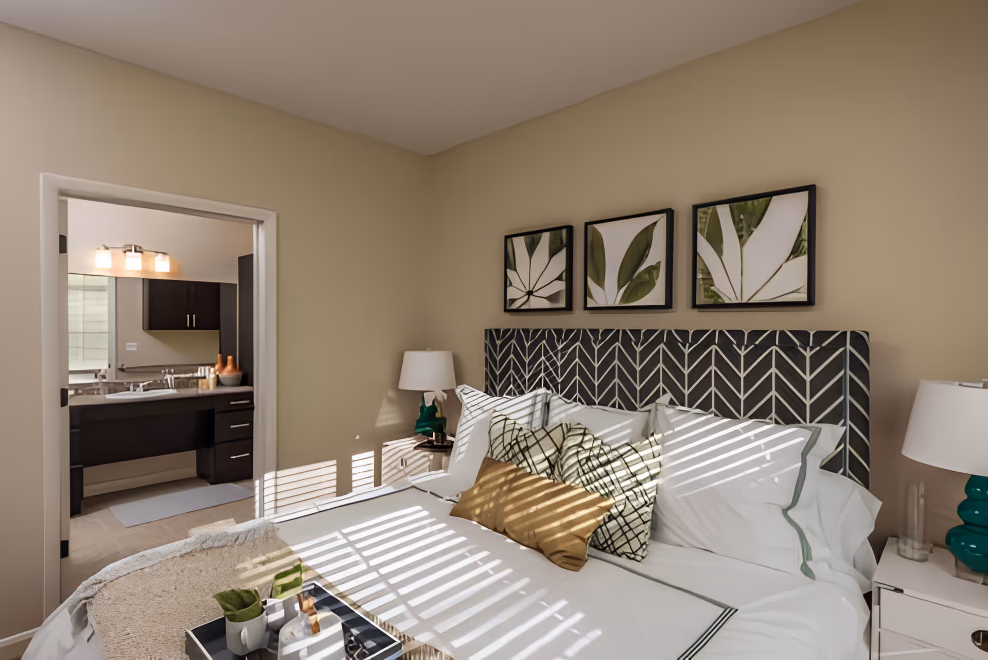 A cozy bedroom with a neatly made bed featuring multiple pillows and a patterned headboard. Three framed botanical prints hang above the bed. A bedside table with a green lamp and a glass is visible. An open doorway leads to a bathroom with a sink, mirror, and dark cabinetry.