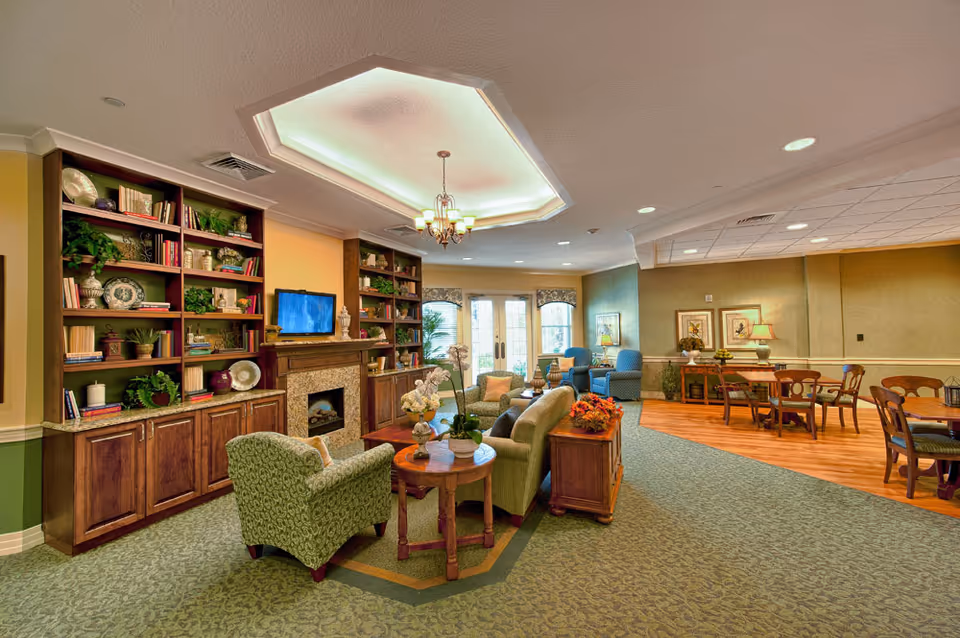 A cozy senior living facility common area with a green patterned armchair, a beige sofa, wooden side tables with flower arrangements, and a fireplace with built-in bookshelves filled with books and decorative items. There is a flat-screen TV above the fireplace. In the background, there are blue armchairs near large windows with curtains and a dining area with wooden tables and chairs. The room has a mix of carpet and wood flooring and a ceiling light fixture.