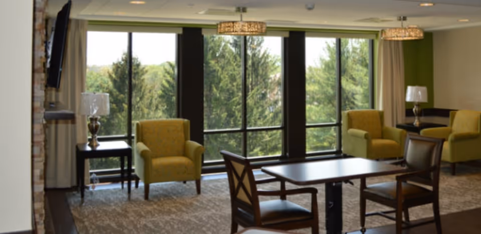 A bright and spacious living room area with large floor-to-ceiling windows showing green trees outside. The room features several comfortable armchairs in a light green color, a wooden table with chairs, two table lamps on side tables, and modern ceiling light fixtures.