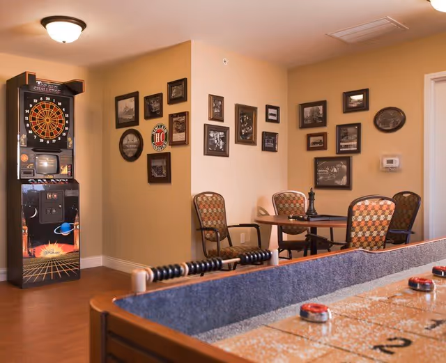 A cozy game room with a dartboard arcade machine against the wall, a small table with four patterned chairs, and a shuffleboard table in the foreground. The walls are decorated with various framed pictures and artwork.